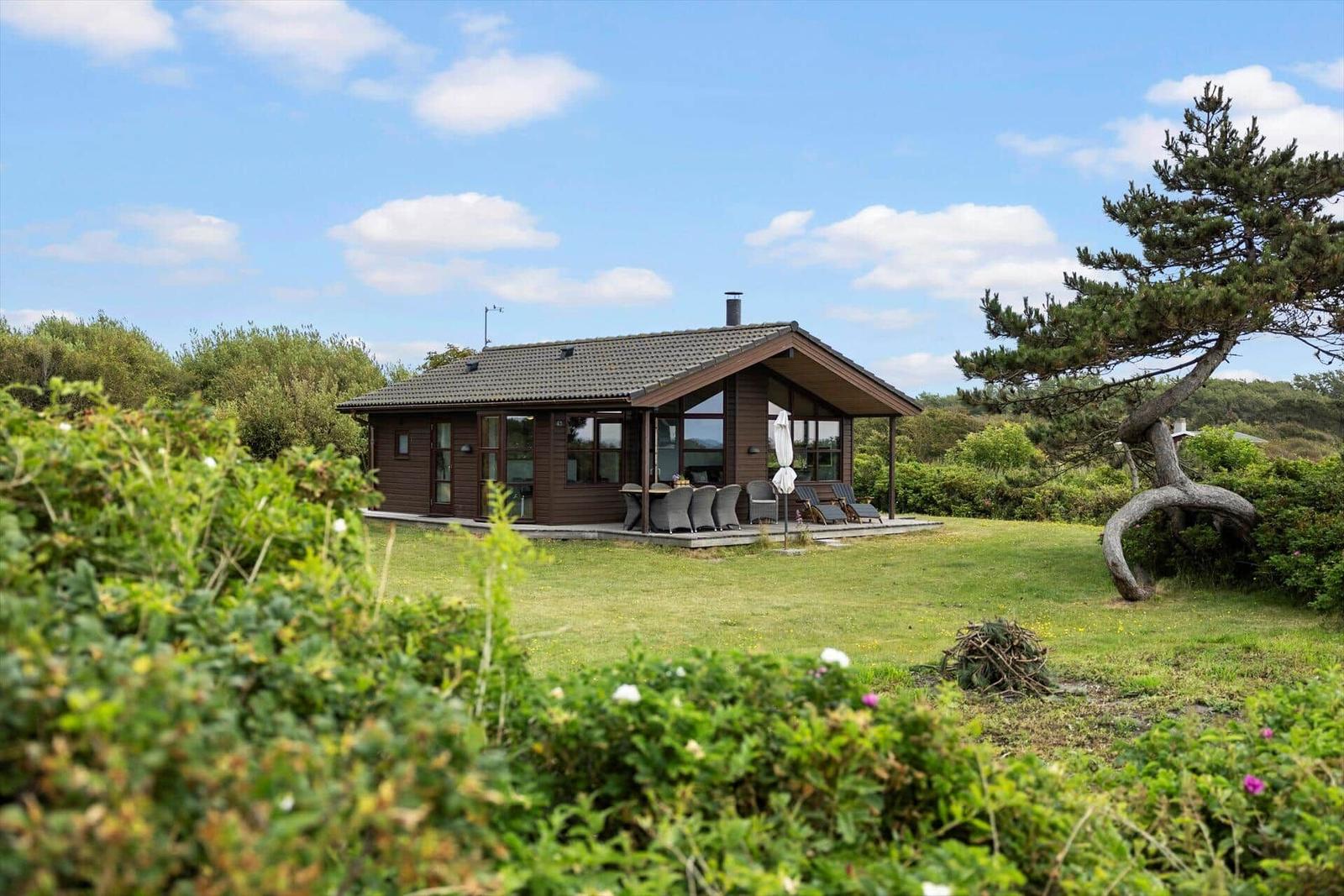 Holzhaus mit Terrasse, Garten und Blick auf Bäume unter blauem Himmel.