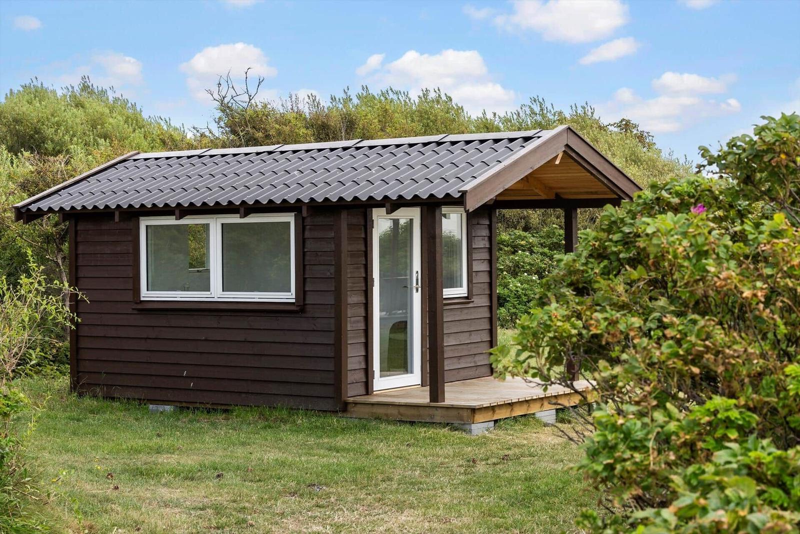 Holzchalet mit Dachüberstand, Fenster und Tür. Umgeben von Gras und Sträuchern.