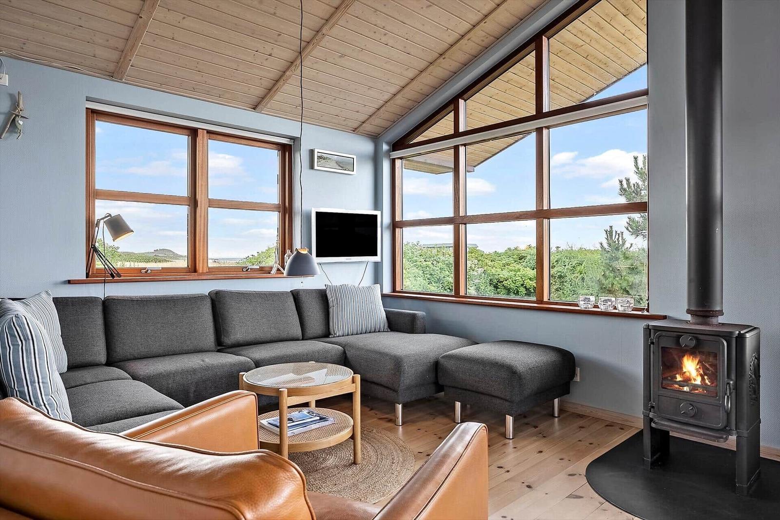 Ein Wohnzimmer mit grauem Sofa, Holzfußboden und Kamin. Große Fenster bieten Blick auf die Landschaft.