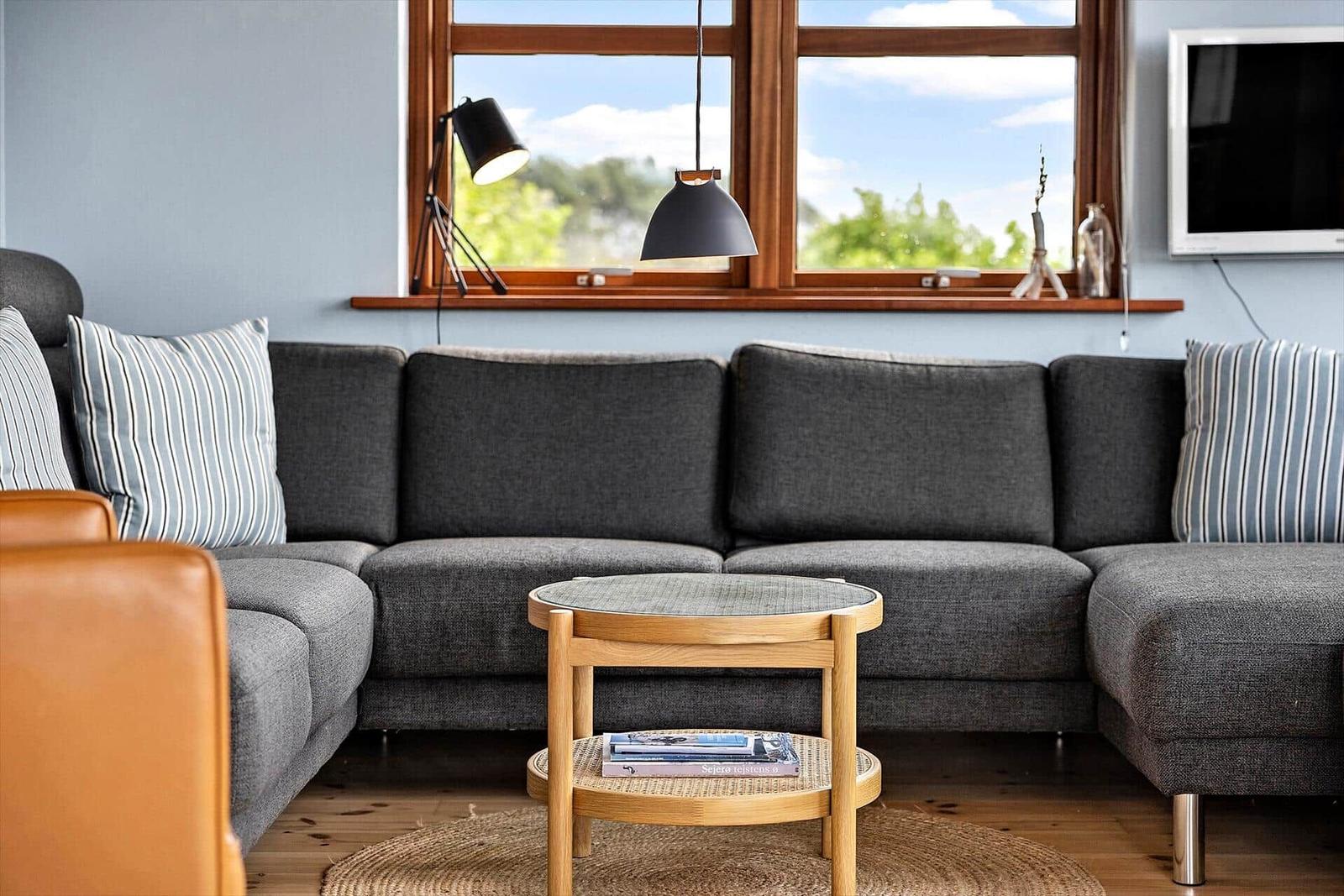 Ein Wohnzimmer mit grauem Sofa, Holztisch und Fenster mit Blick auf Grün.