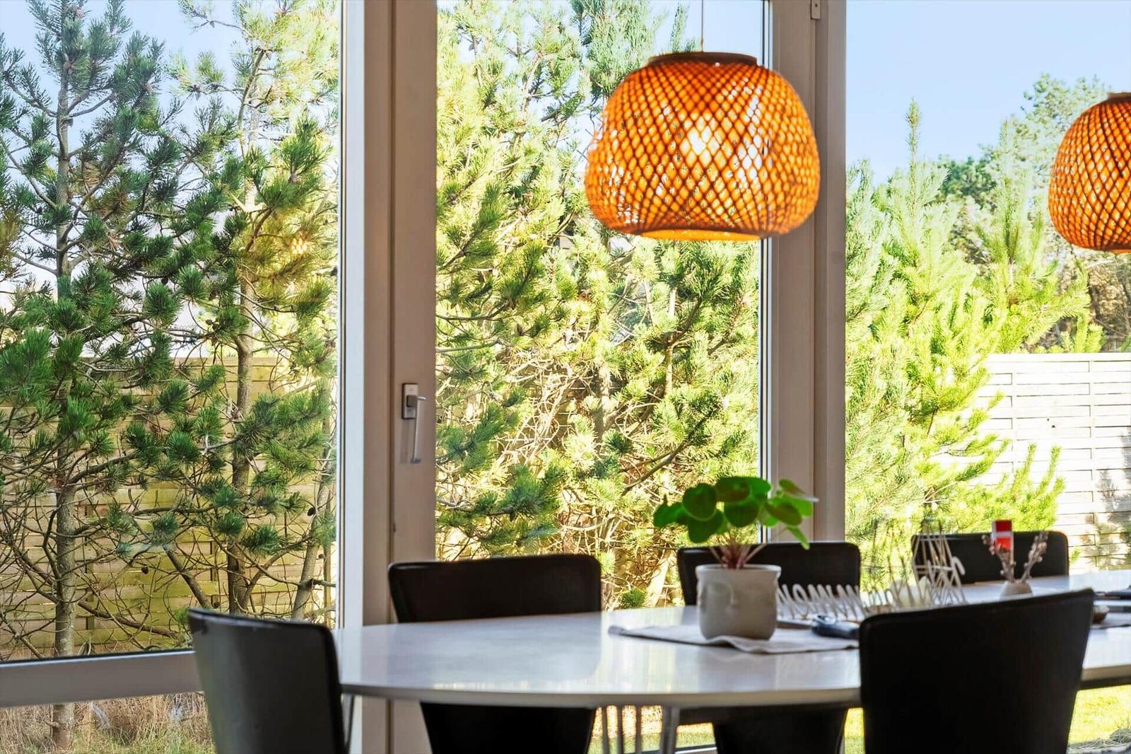 Dining area with table, chairs, and pendant lamp. View of green trees through large windows.