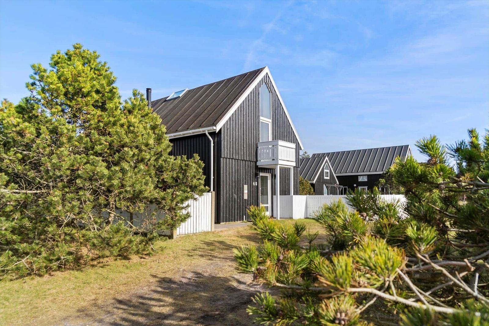 Black wooden house with balcony and skylight, surrounded by pine trees and white fence.