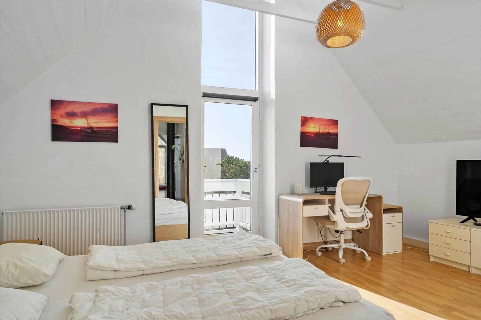 Bedroom with double bed, desk, and TV. Large windows and wooden floor.