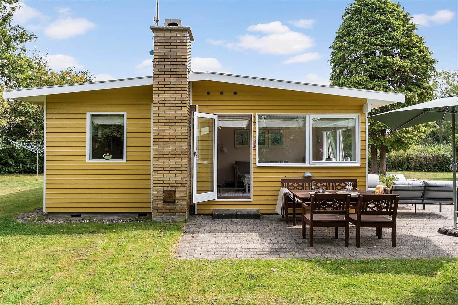 Gelbes Haus mit Terrasse, Tisch, Stühlen und Garten. Durchgangstür zu Wohnbereich.