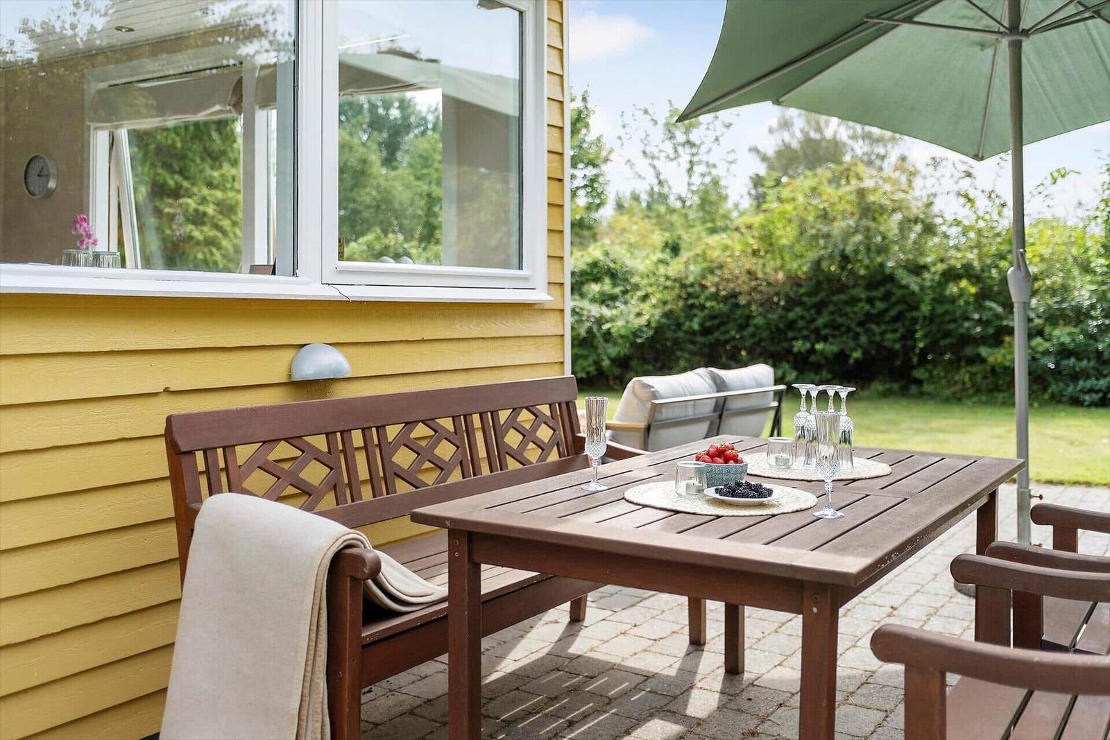 Terrasse mit Tisch, Bänken und Sonnenschirm. Hintergrund: grüner Garten und gelbes Haus.