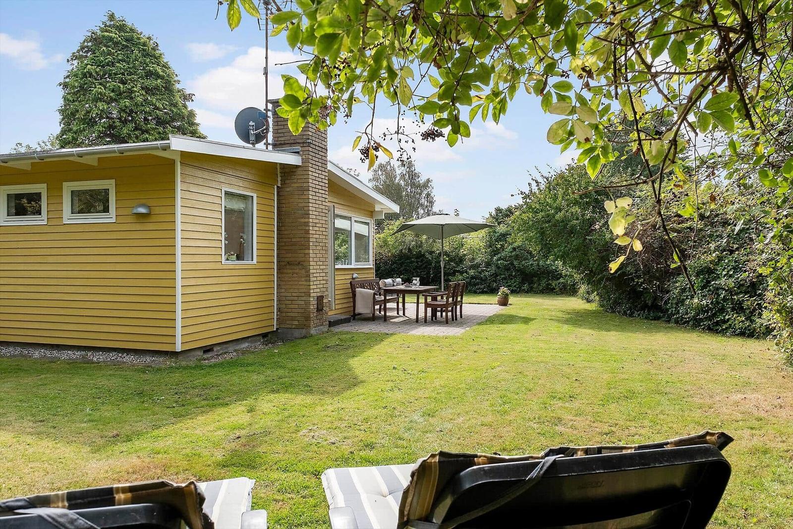 Gelbes Haus mit Garten, Terrasse und Sonnenschirm. Liegestühle im Vordergrund.