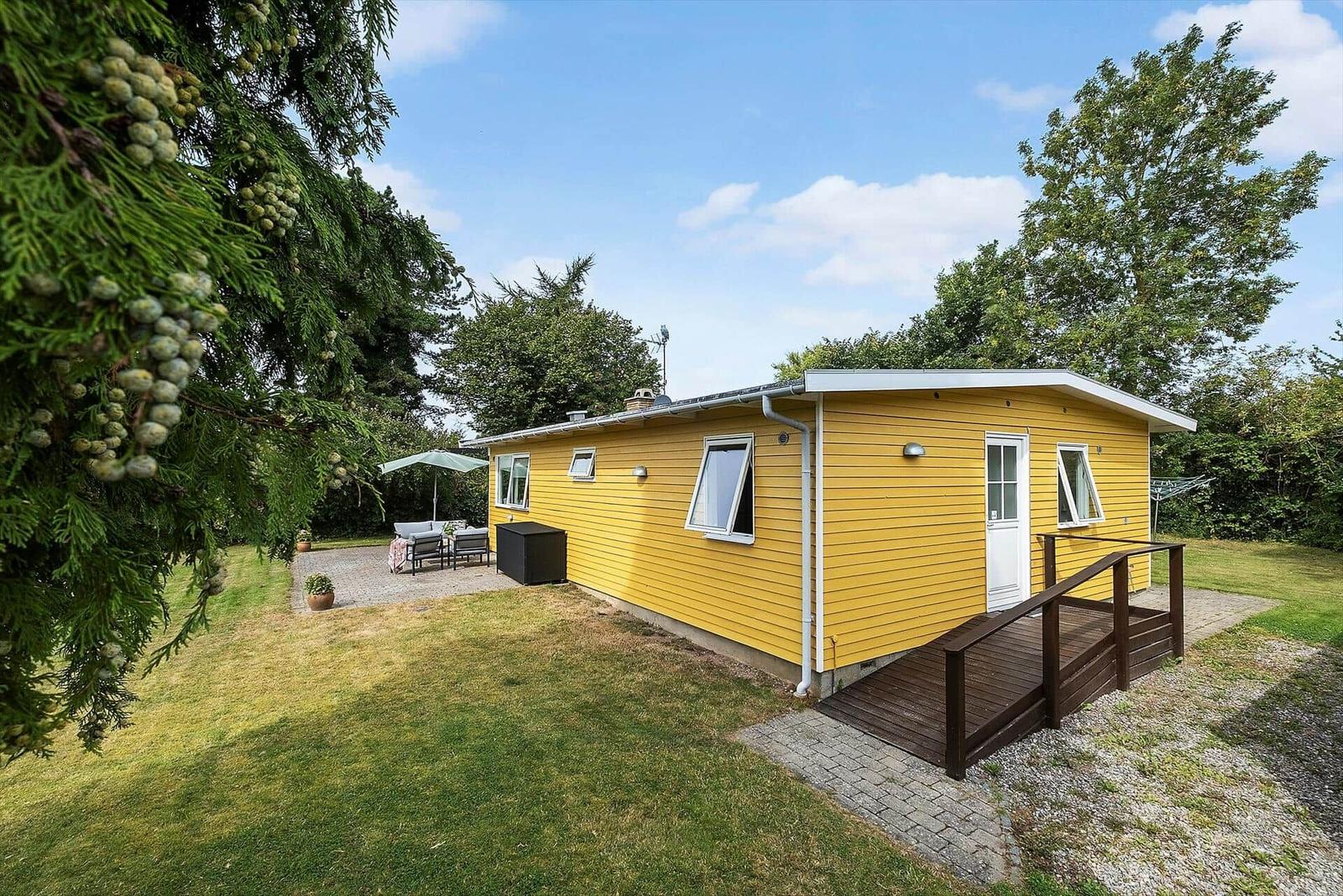 Gelbes Ferienhaus mit Terrasse und Garten unter Bäumen.