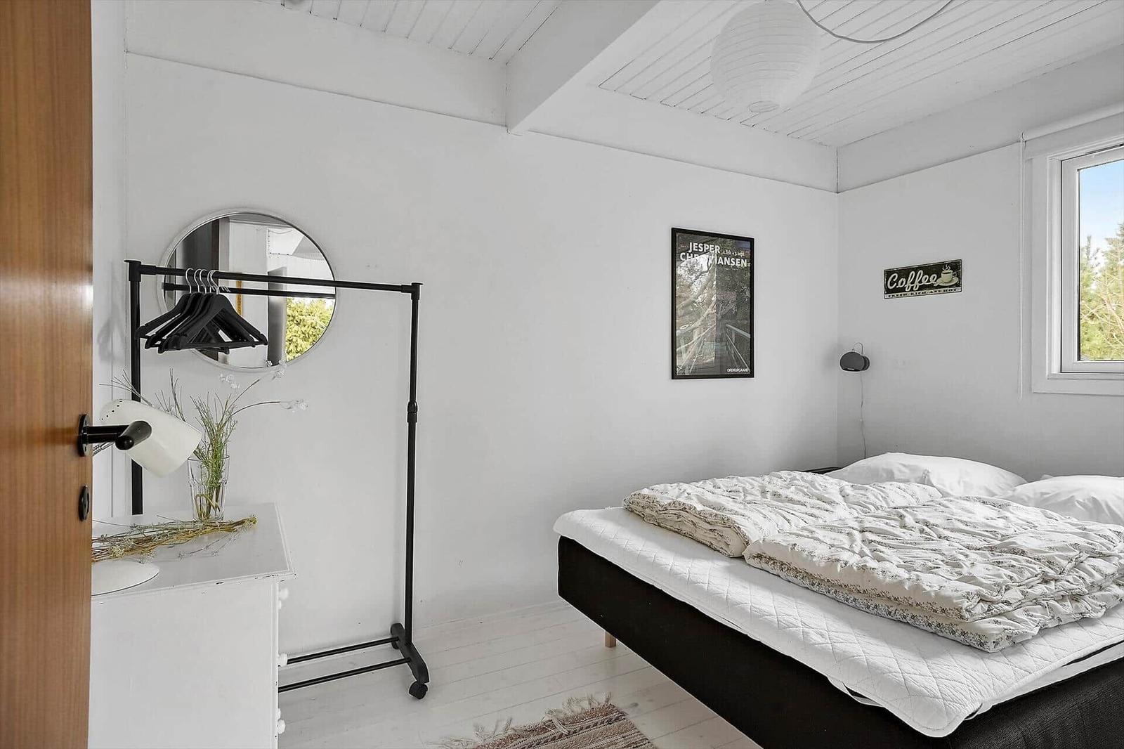 Schlafzimmer mit Bett, Kleiderständer und Fenster. Wände weiß, Boden hell.