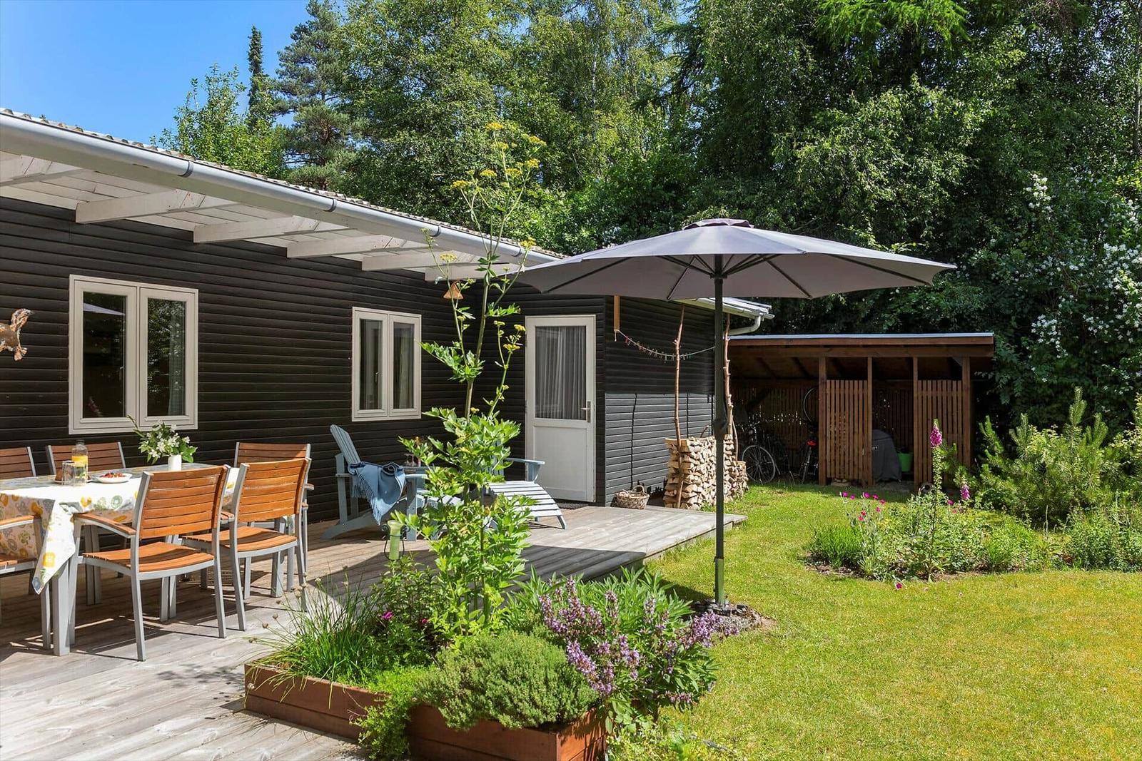 Haus mit Terrasse, Garten und Sonnenschirm. Tisch mit Stühlen und Pflanzen.