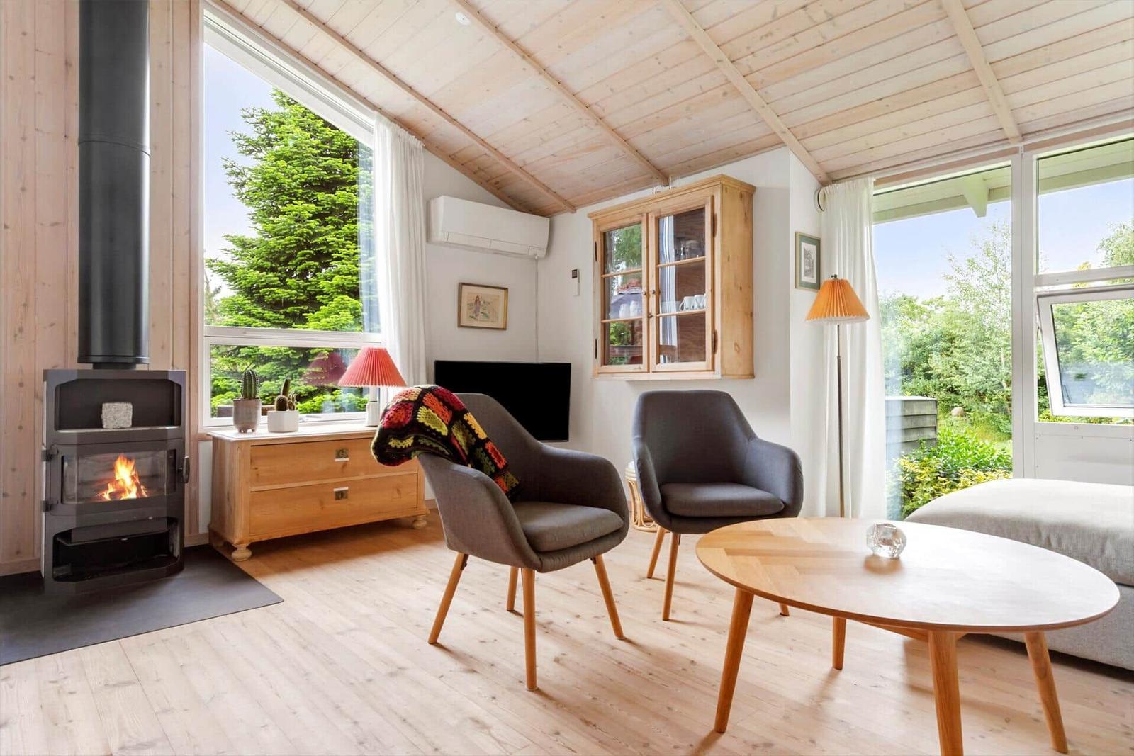 Wohnraum mit Kamin, zwei Sesseln und Holztisch. Große Fenster zeigen grüne Bäume.