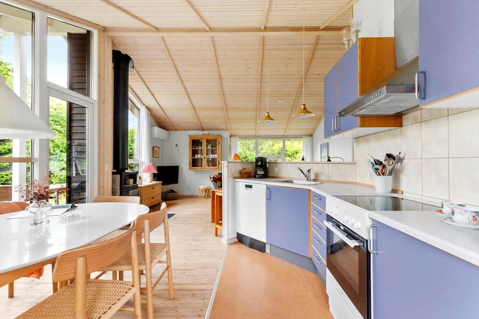 Küche mit blauen Schränken und Esstisch. Holzdecke und große Fenster.