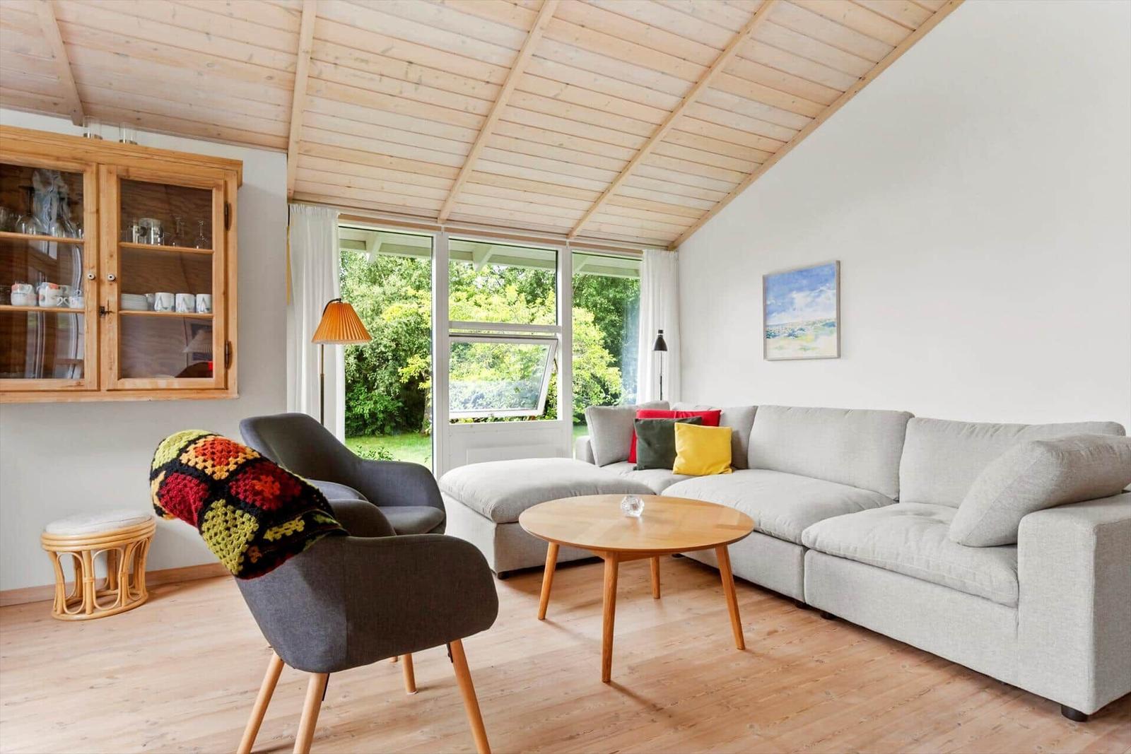 Wohnzimmer mit Sofa, Sessel und Holzdecke. Fenster blicken auf grüne Außenanlage.