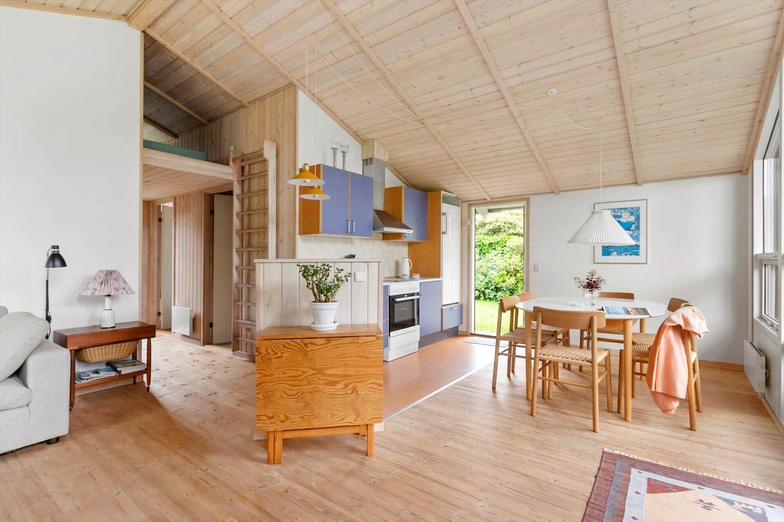 Wohn- und Essbereich mit Küche unter Holzdecke. Tisch mit Stühlen, Sofa und Pflanze.