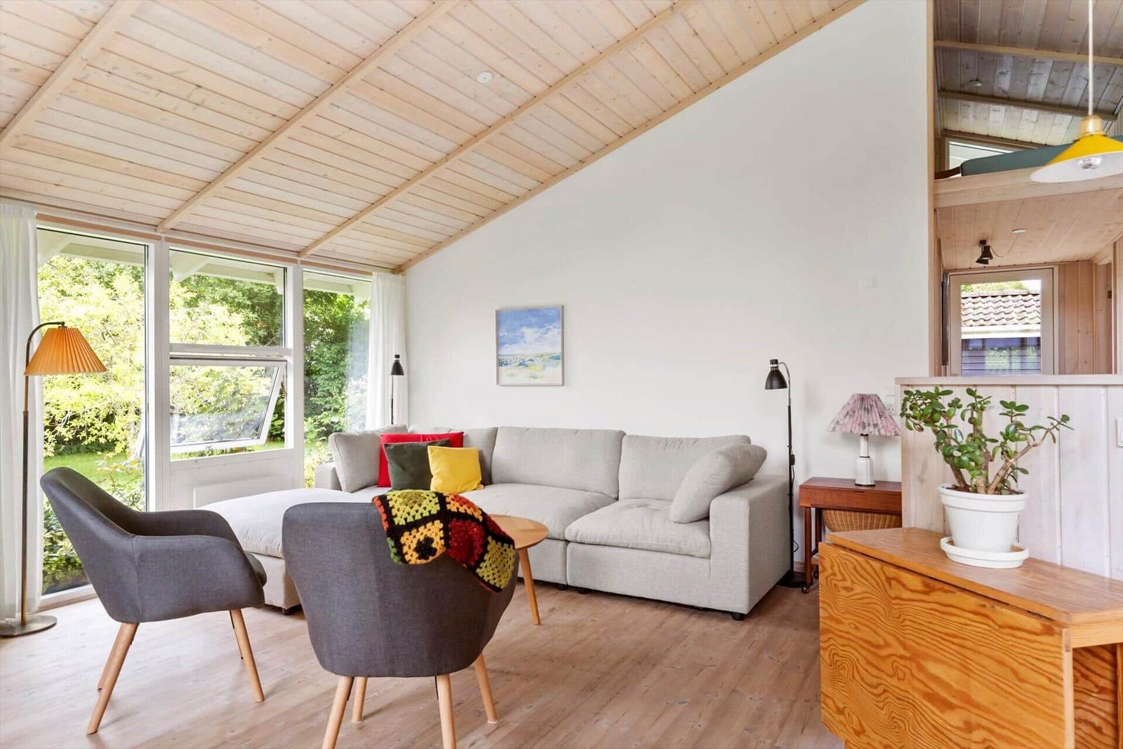 Wohnzimmer mit Sofa, zwei Stühlen und Holzboden. Große Fenster und Holzdecke.