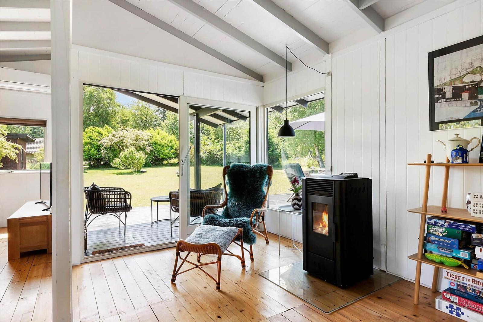 Raum mit Holzboden, Kamin und Blick auf Garten. Sitzgelegenheit und Bücherregal sichtbar.