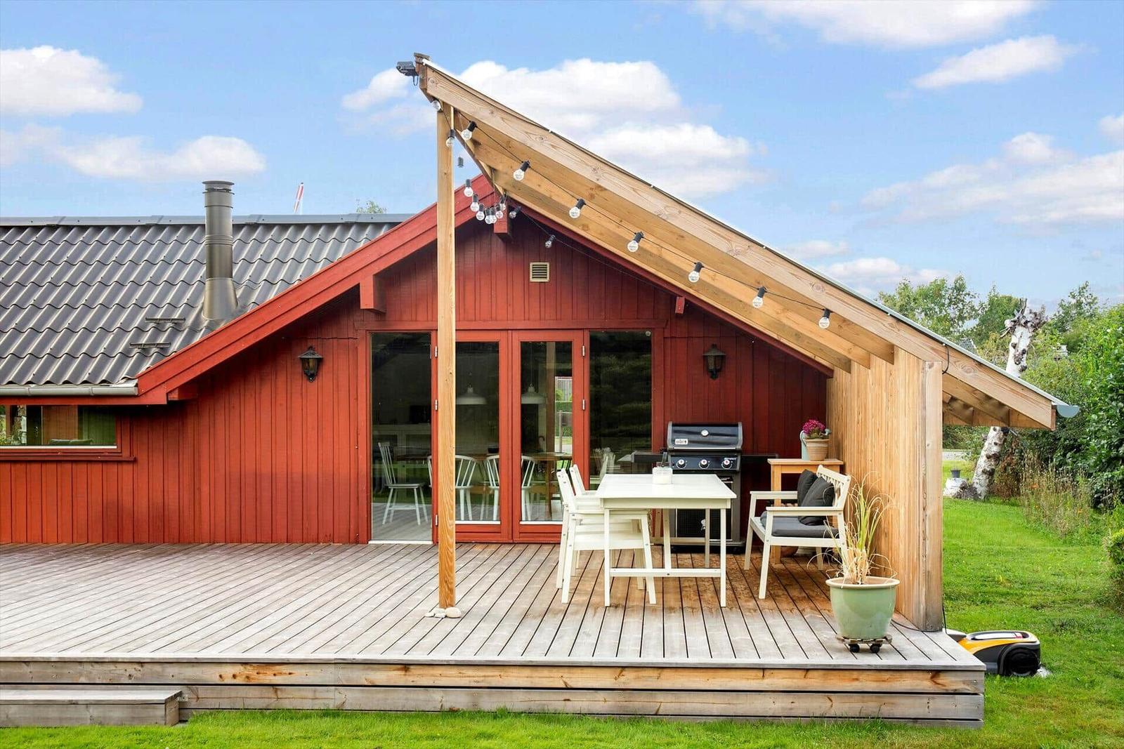 Rotes Holzhaus mit Terrasse, Grill und Sitzgelegenheiten unter einem Dach.