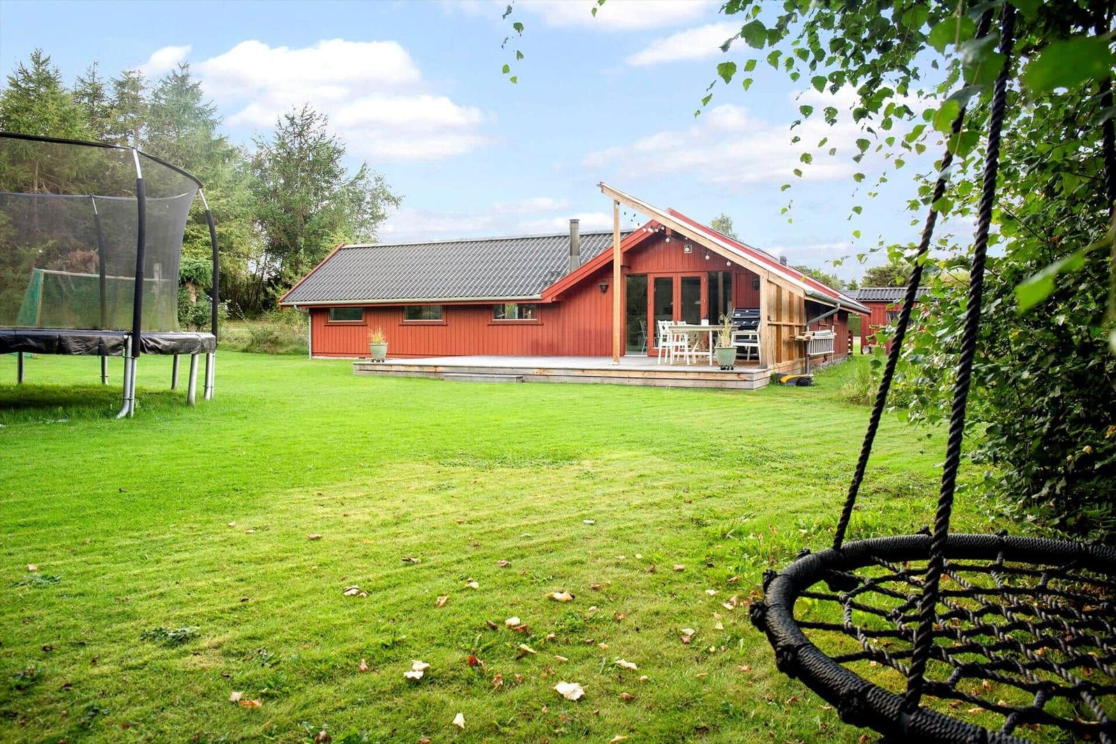 Roter Holzausstattungshaus mit Terrasse, Trampolin und Schaukel im Garten.