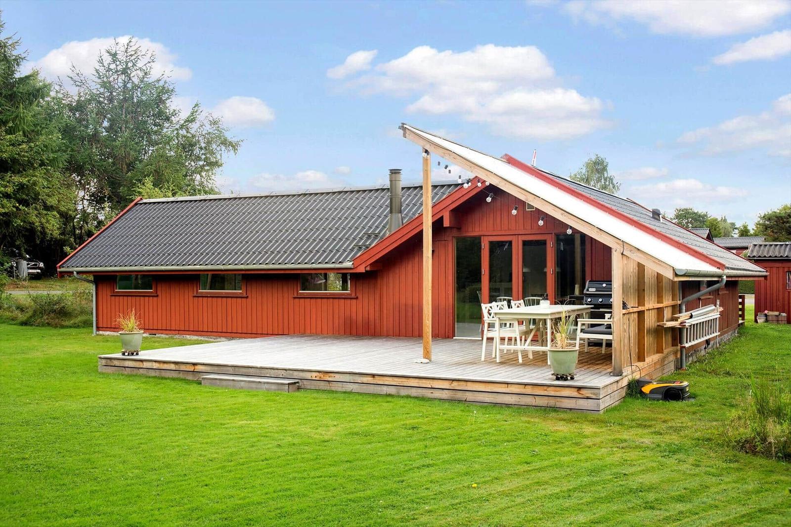 Rotes Ferienhaus mit Holzterrasse und Grillplatz im Grünen.