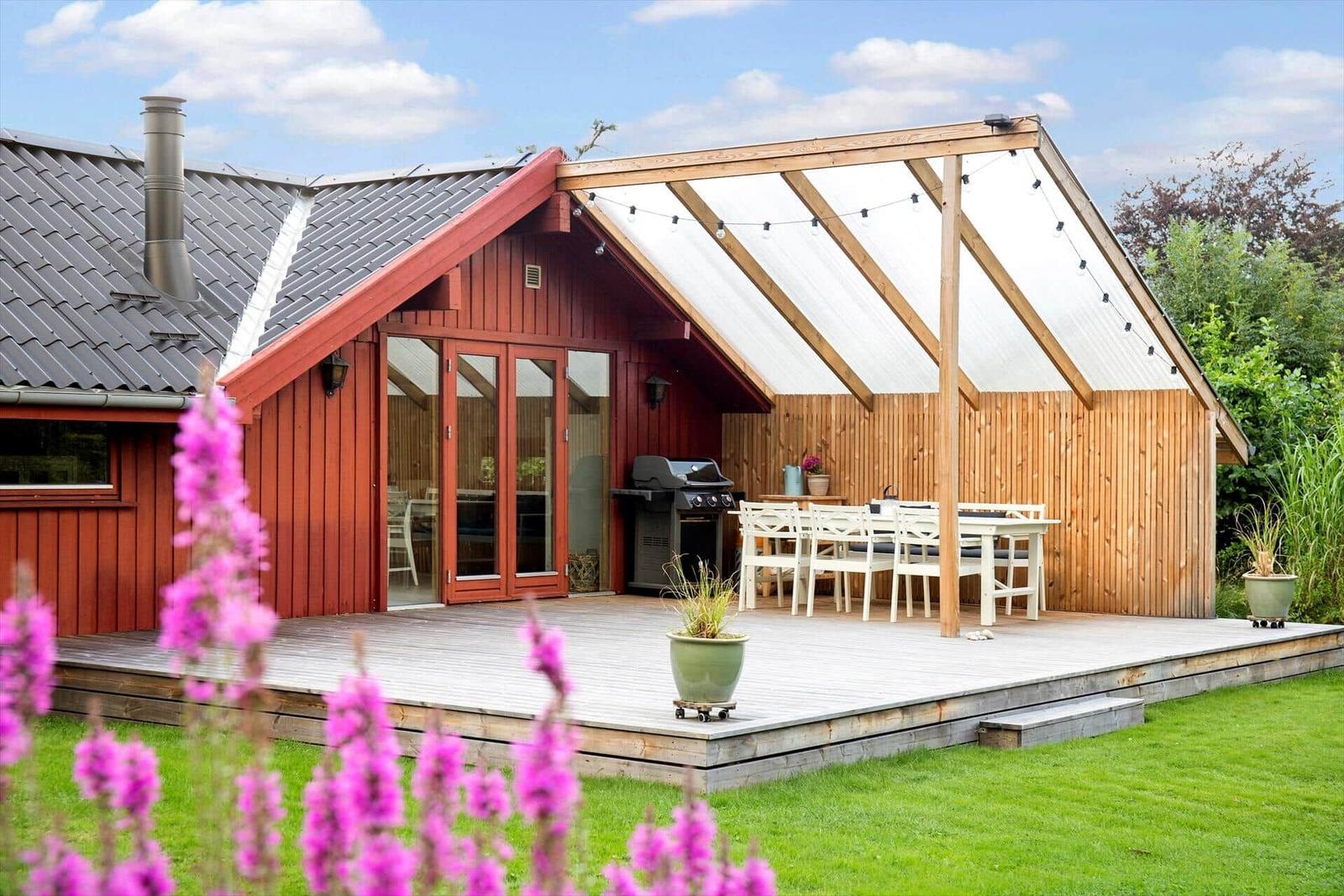 Roter Holzhaus mit Terrasse, Grill und weißem Tischset im Garten.