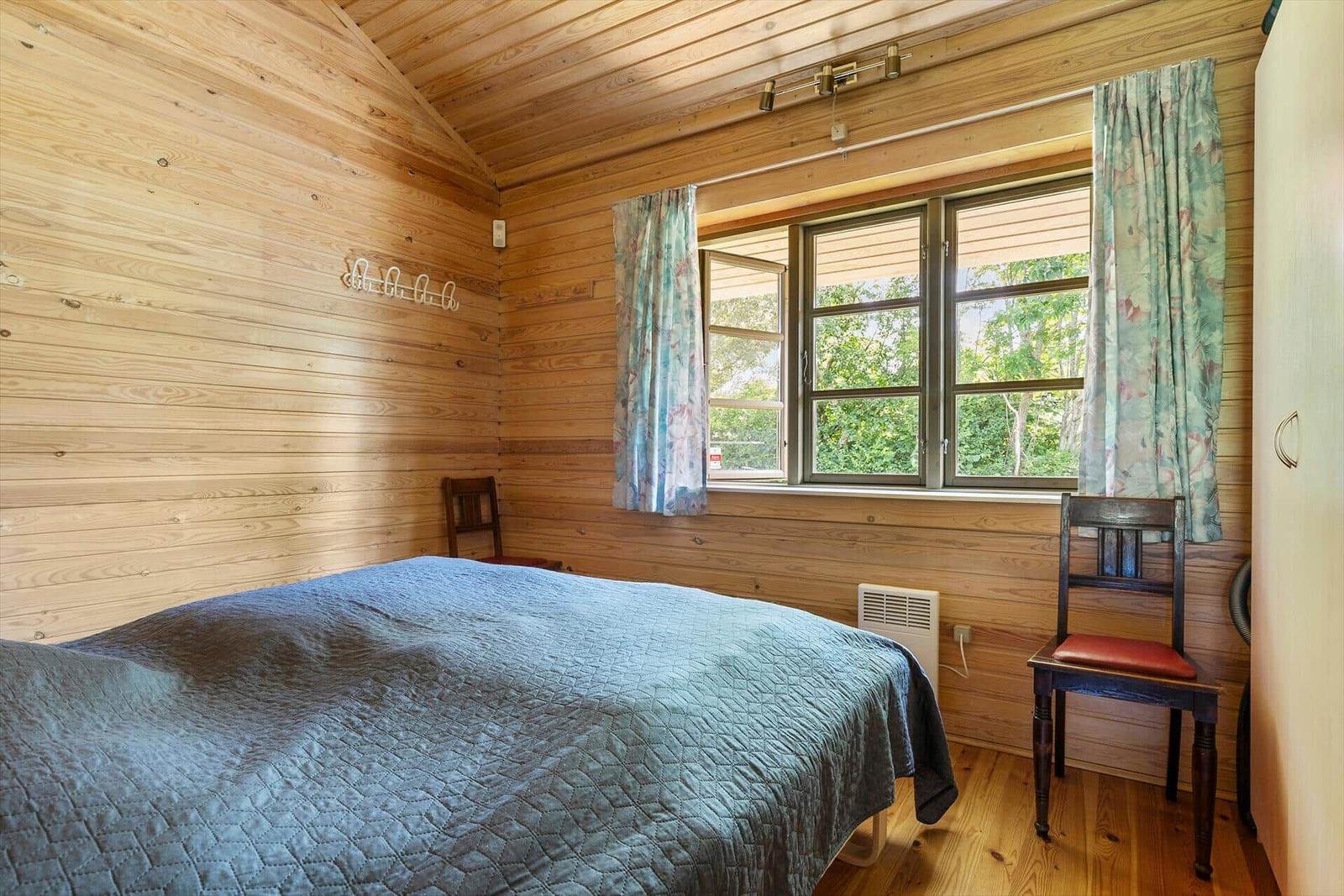 Schlafzimmer mit Holzwand, Bett, Stuhl und Fenster mit Vorhängen.