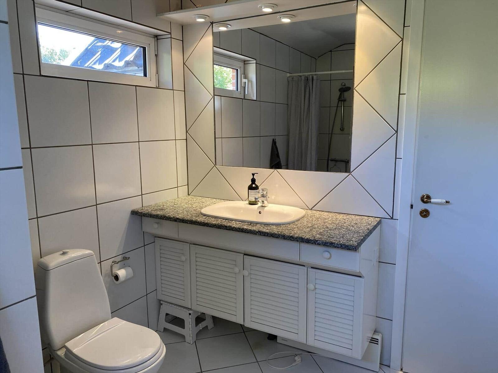 Badezimmer mit Waschbecken, Spiegel und Toilette. Fliesenwand und Fenster sichtbar.