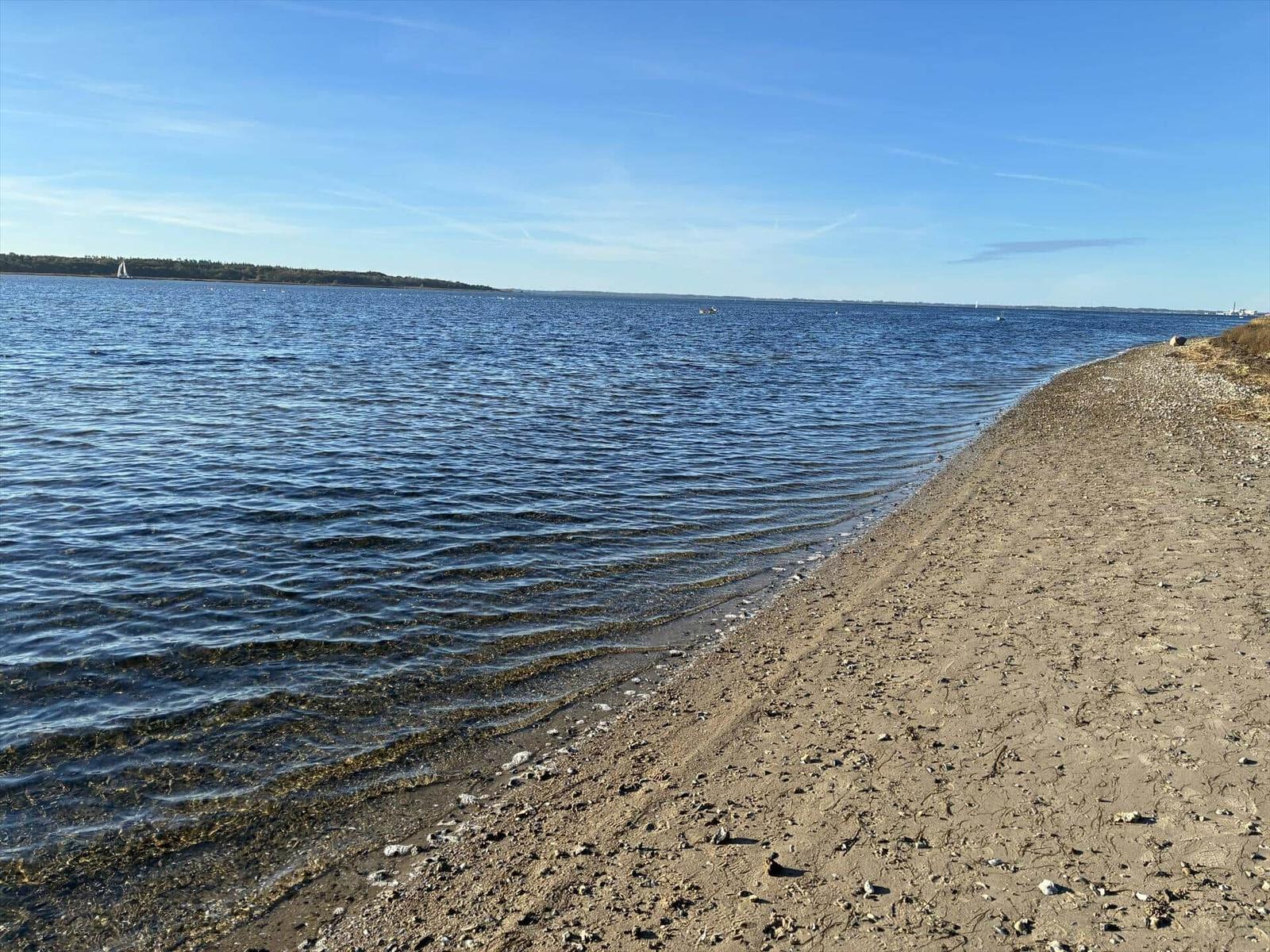 Strand mit Sand und Steinen am Wasser, Boot im Hintergrund unter blauem Himmel.