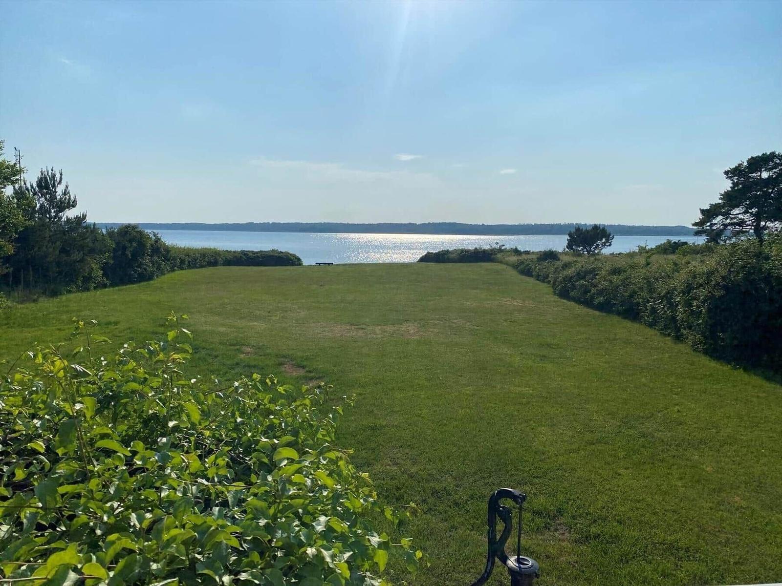 Grüner Rasenhang mit Blick auf ruhiges Wasser und Bäume im Hintergrund.