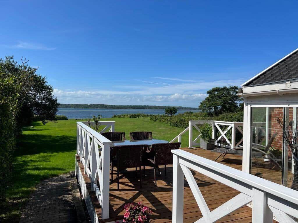 Terrasse mit Tisch und Stühlen, Blick auf ruhiges Wasser und grünes Gelände.