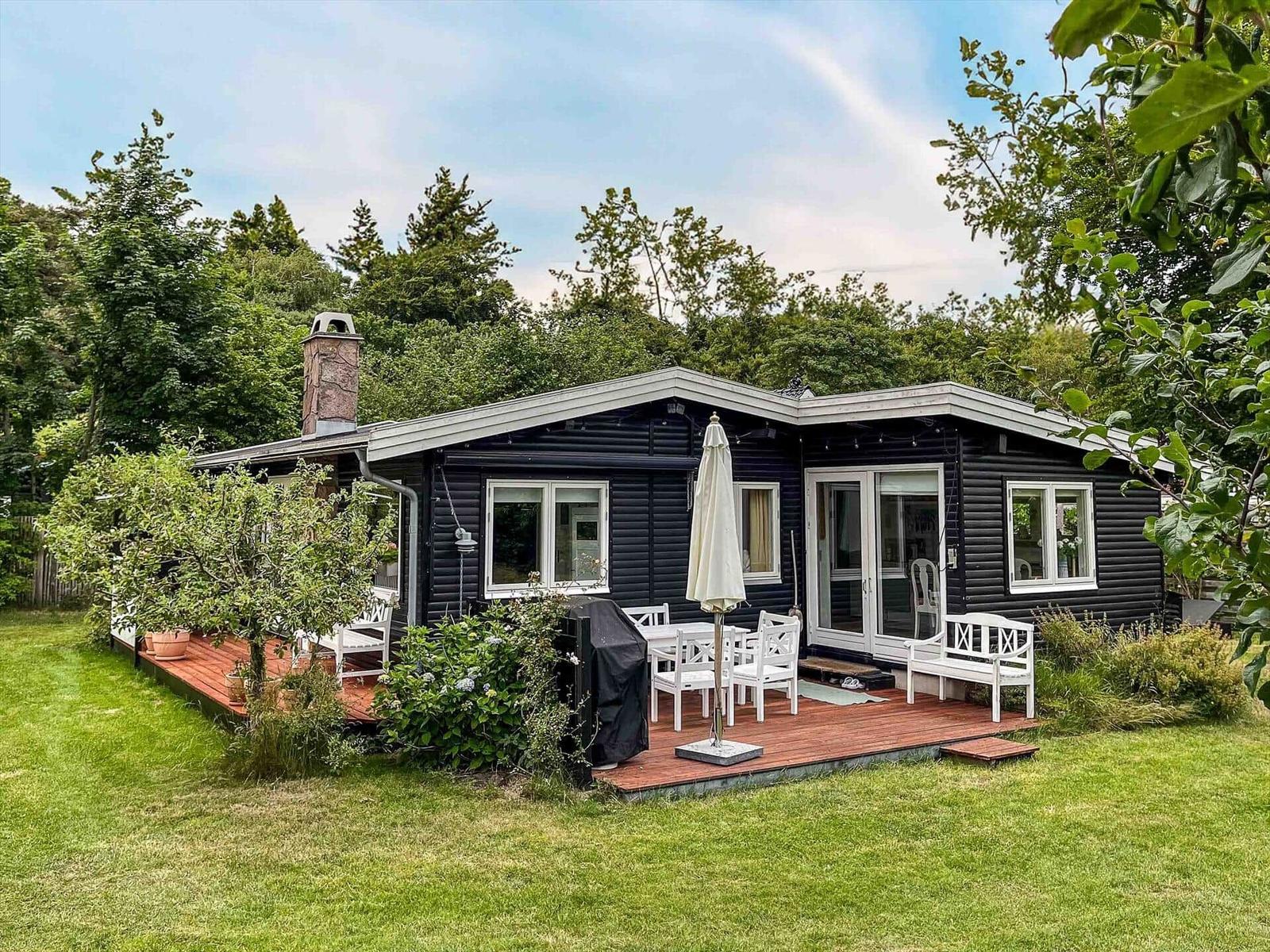 Schwarzes Holzhaus mit Terrasse, weißen Stühlen und Grill. Umgeben von Gras und Bäumen.