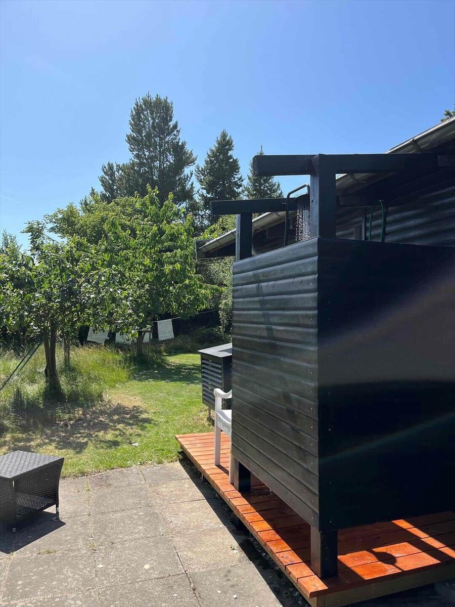 Schwarzer Holzschuppen mit Terrasse, Garten und Bäumen im Hintergrund.