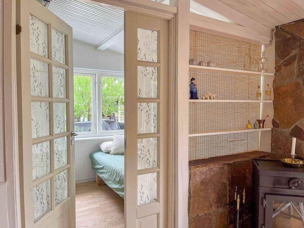 Durchgehende Tür führt in ein Schlafzimmer mit Bett und Fenster. Nebenher steht ein Kamin mit Regalen.