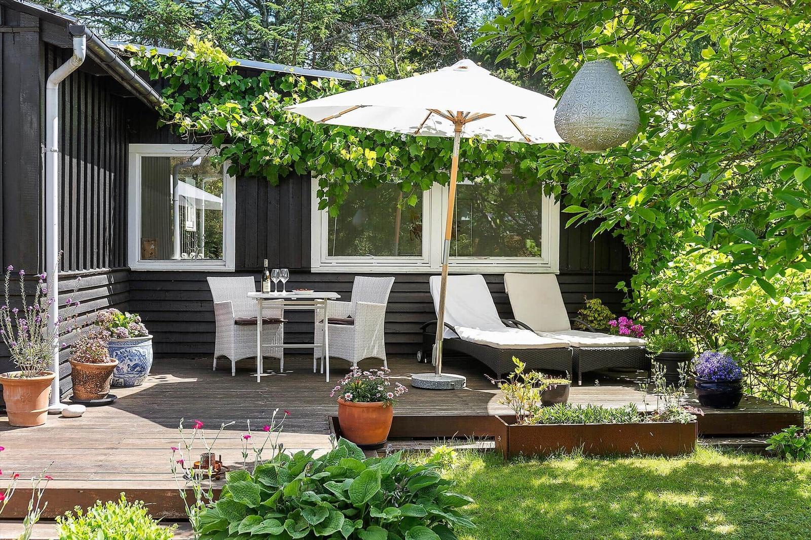Terrasse mit Tisch, Stühlen, Liegen und Sonnenschirm. Umgeben von Pflanzen und Grün.