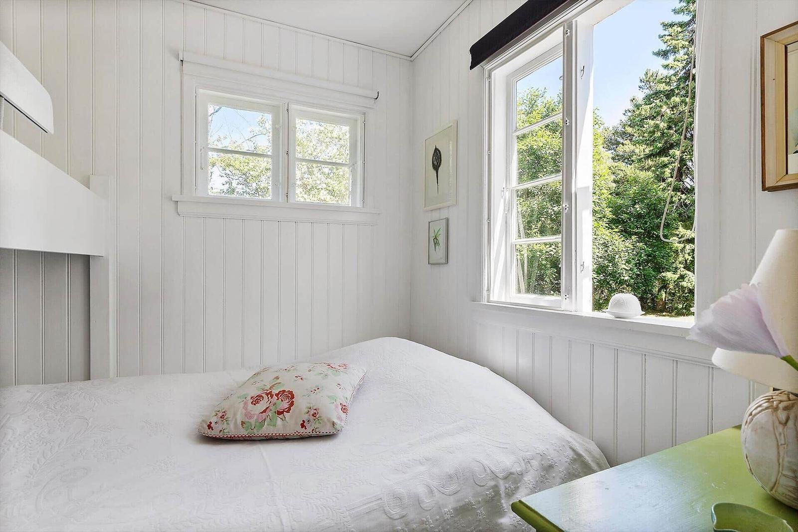 Schlafzimmer mit weißer Wandverkleidung, Bett und Fenster mit Blick auf Bäume.