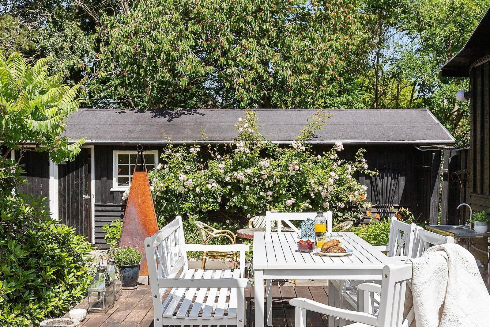 Hinterhof mit weißem Tisch, Stühlen und Blumenhecken. Schwarzes Holzhaus im Hintergrund.