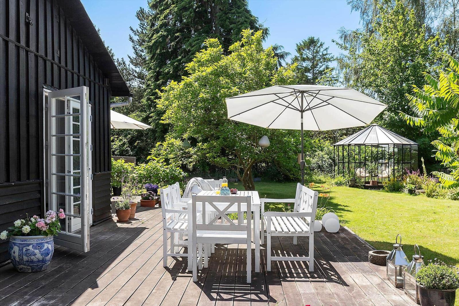 Terrasse mit weißem Tisch und Stühlen unter Sonnenschirm. Grünflächen und Gewächshaus im Hintergrund.