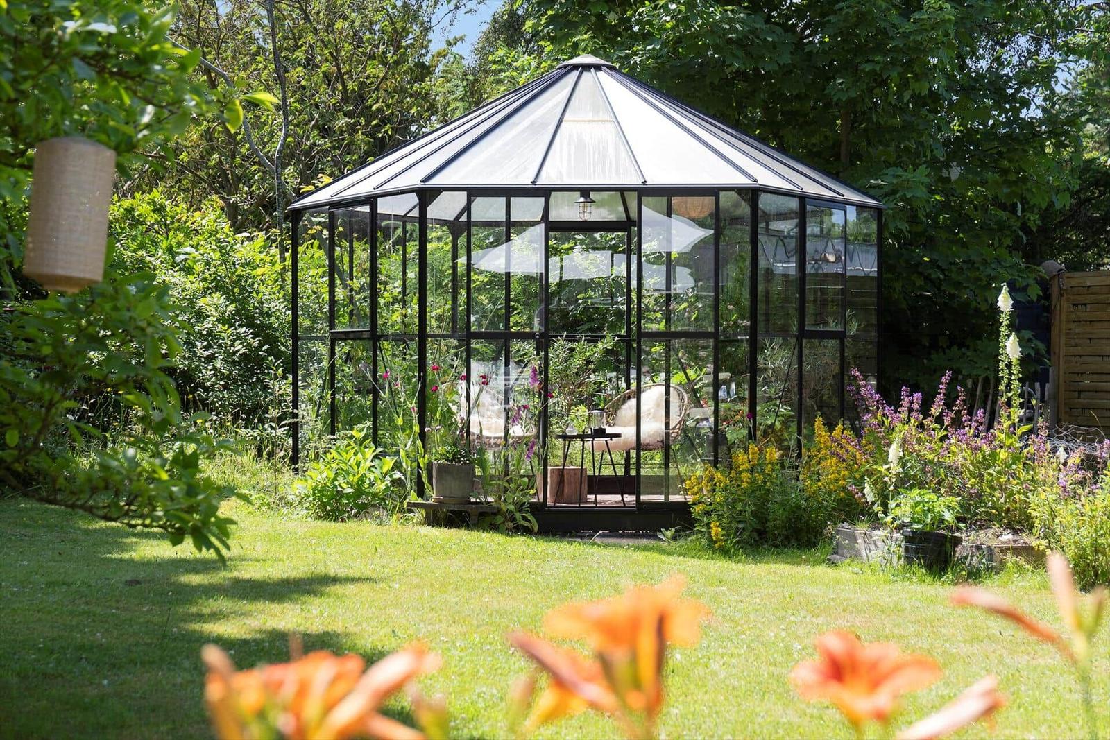 Glasveranda mit Sitzgelegenheit im Garten, umgeben von Pflanzen und Blumen.