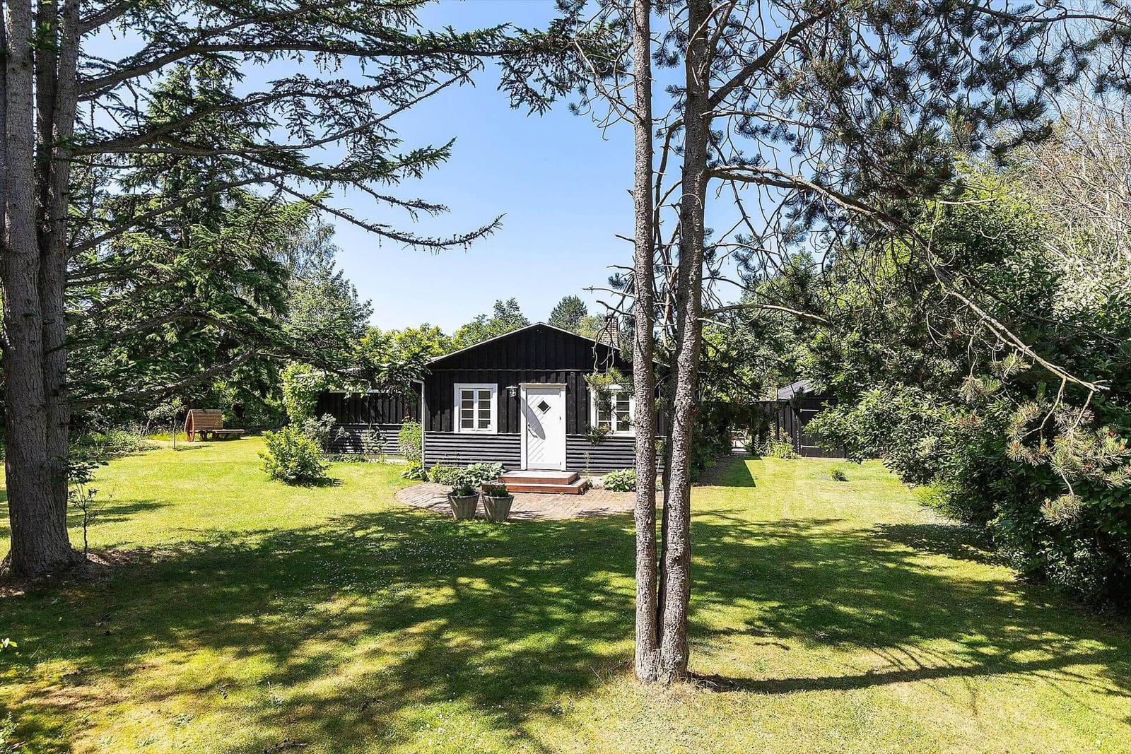 Schwarzes Holzhaus mit weißer Tür, umgeben von Gras und Bäumen.