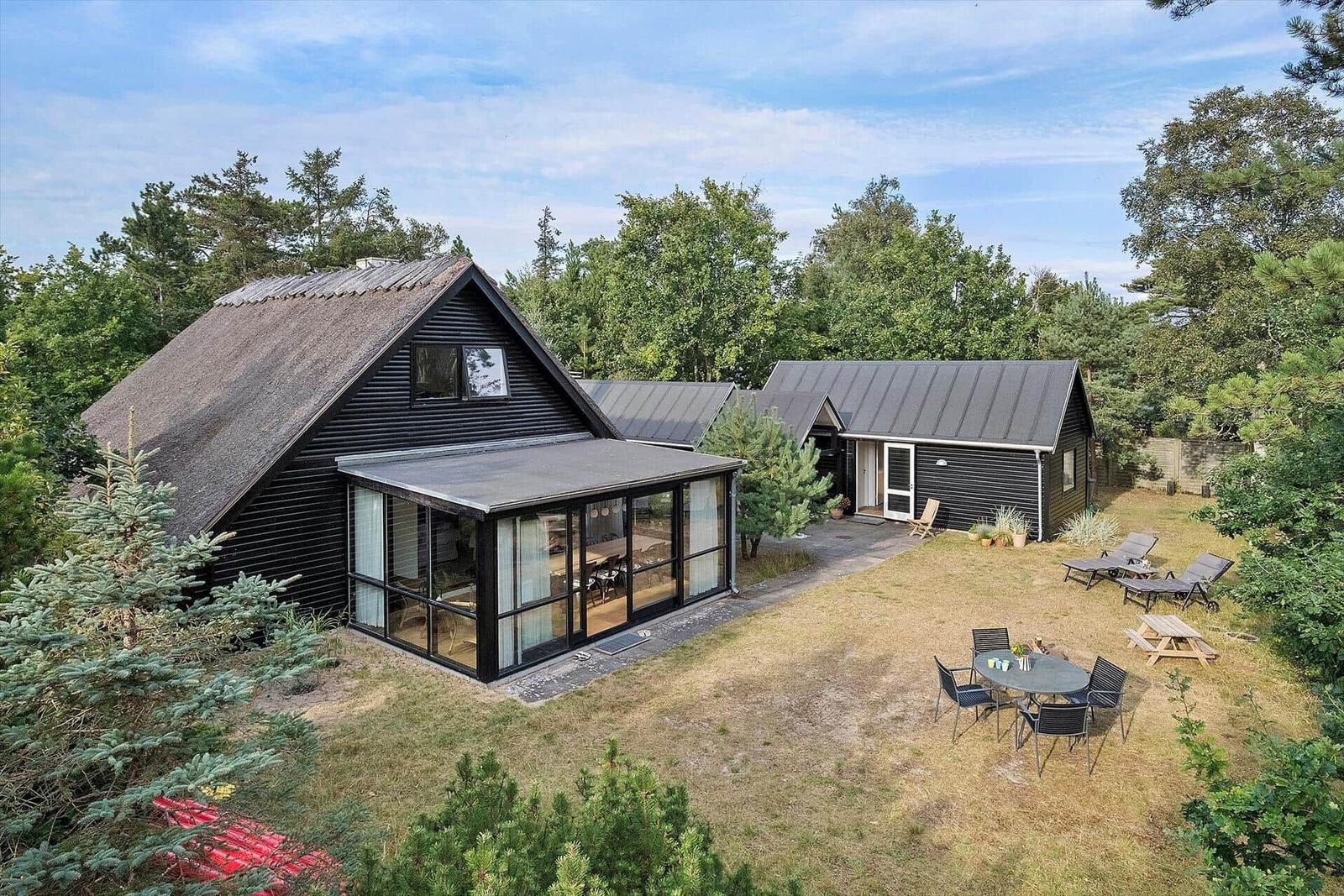 Schwarzes Haus mit Holzfassade und Glasveranda. Garten mit Liegestühlen und Tisch.