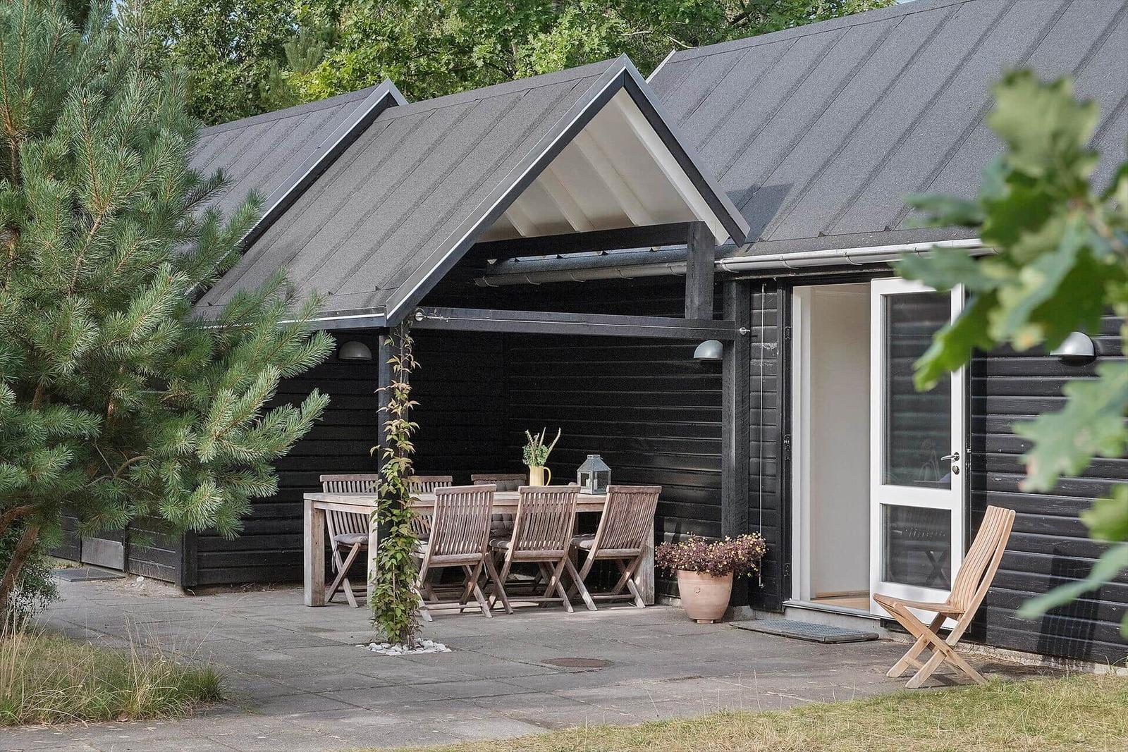 Schwarze Holzhütte mit Terrasse, Tisch und Stühlen. Eingangstür und Pflanzen sind sichtbar.