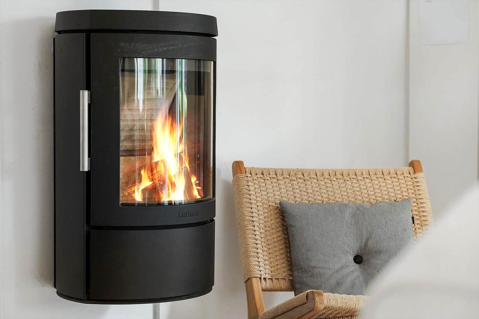 Wandmontierte Kaminofen mit brennendem Feuer neben einem geflochtenen Stuhl mit Kissen.