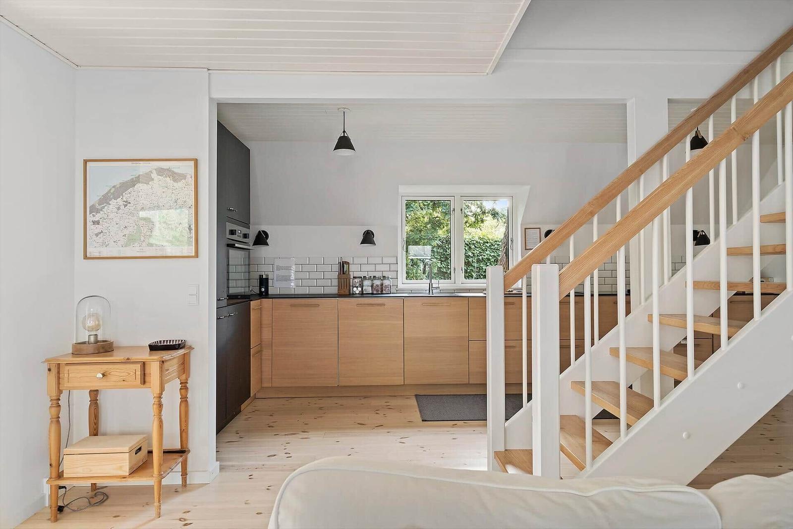 Küche mit Holzschrank und Fenster. Treppe mit Holzgeländer. Holzboden und weiße Wände.