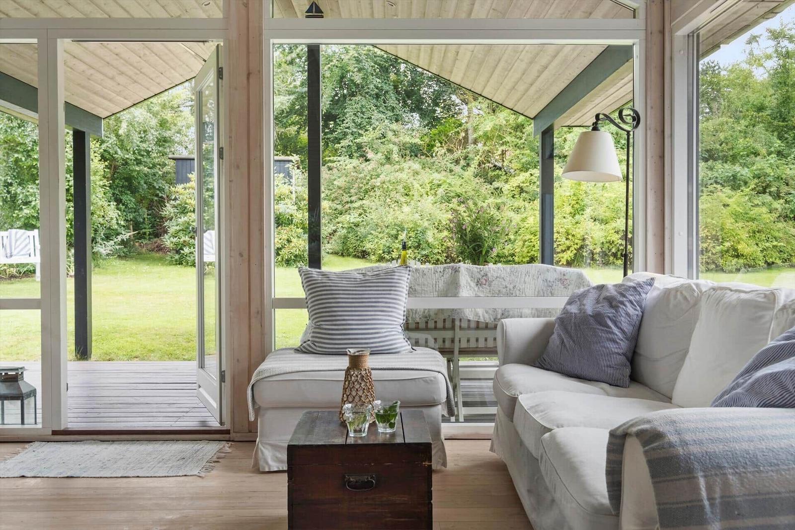 Ein Wohnbereich mit Sofa, Kissen und Tisch. Durchgehende Glasfront zeigt Garten.