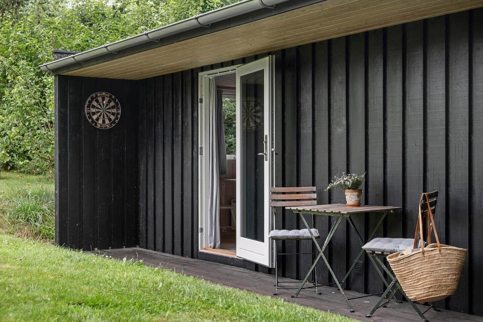 Schwarzer Holzschuppen mit Terrasse, Tisch, Stühlen und Dartscheibe an der Wand.