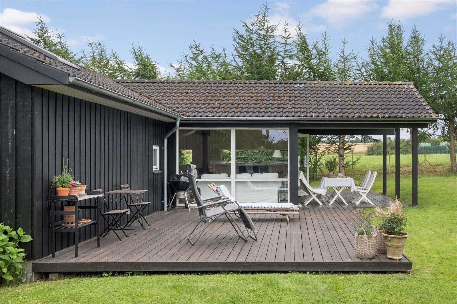 Holzterrasse mit Sitzgelegenheiten und Grill. Blick auf grüne Wiese und Bäume.