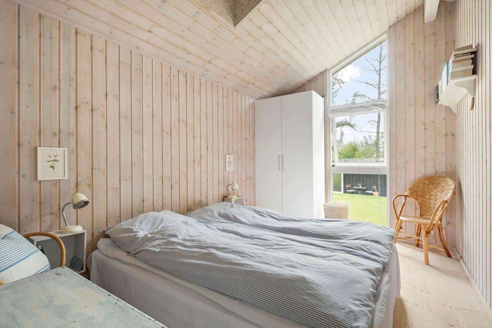 Schlafzimmer mit Holzwand, Bett, Kleiderschrank und Sicht auf Garten.