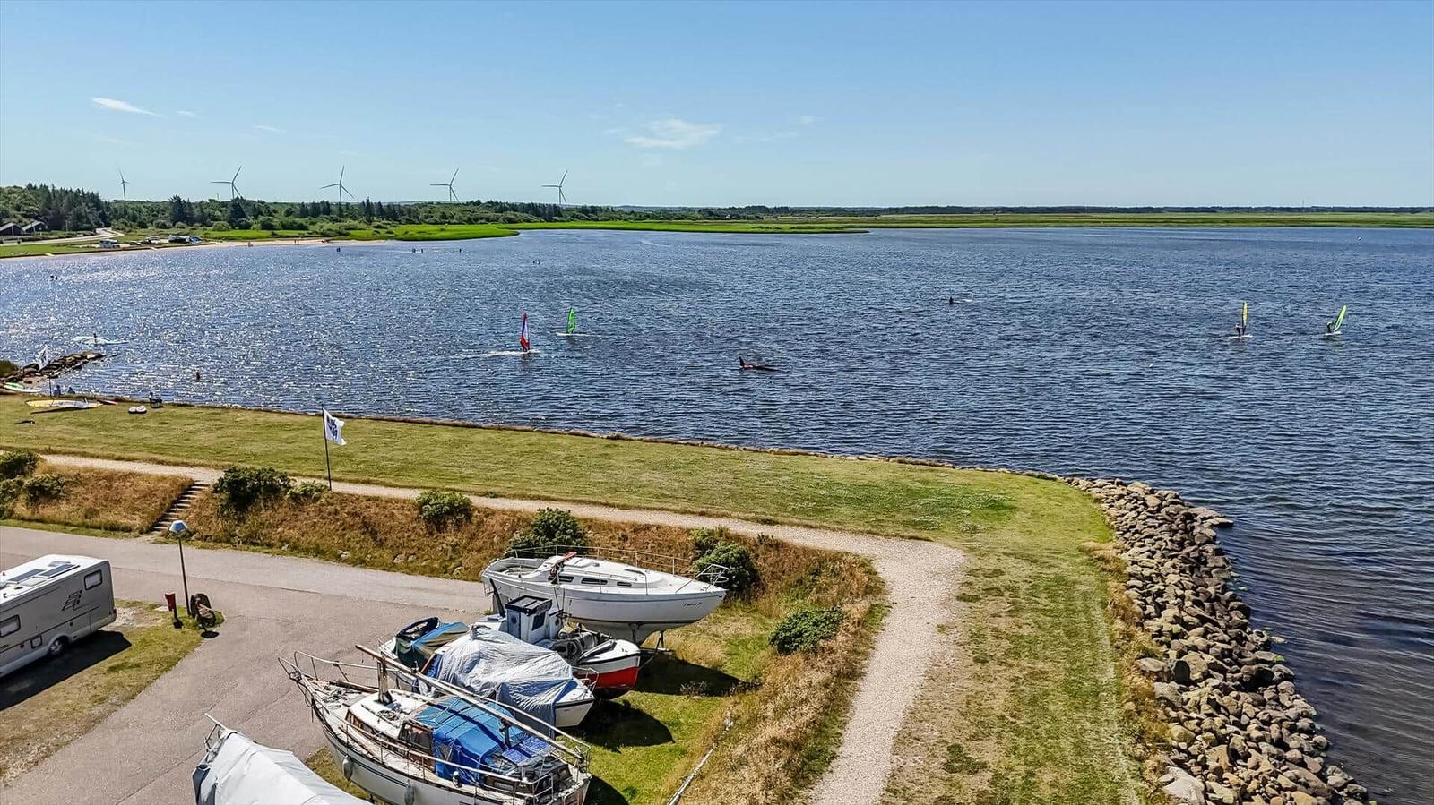 Seeufer mit Booten und Windsurfern unter blauem Himmel.