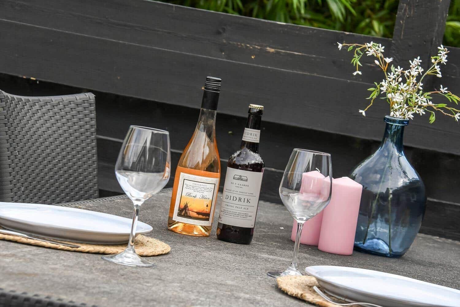 Außenbereich mit Tisch, zwei Flaschen, Gläsern, Kerzen und Blumen in einem Blumentopf.