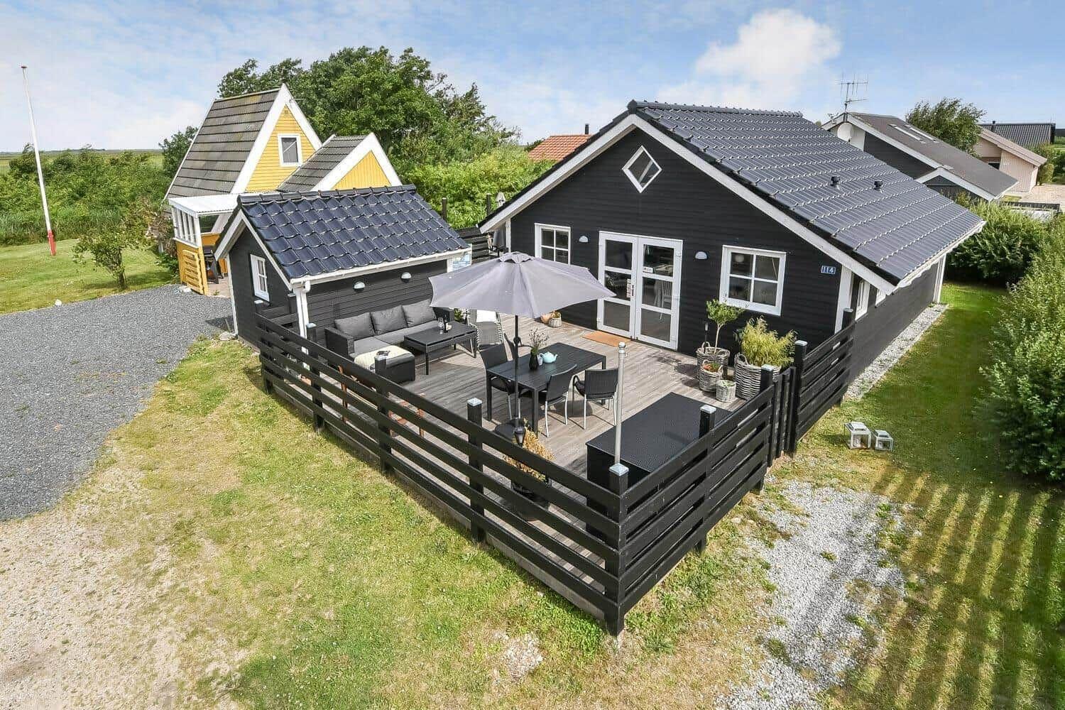 Schwarzes Ferienhaus mit Terrasse, Sitzgruppe und Sonnenschutz. Grün umgeben.