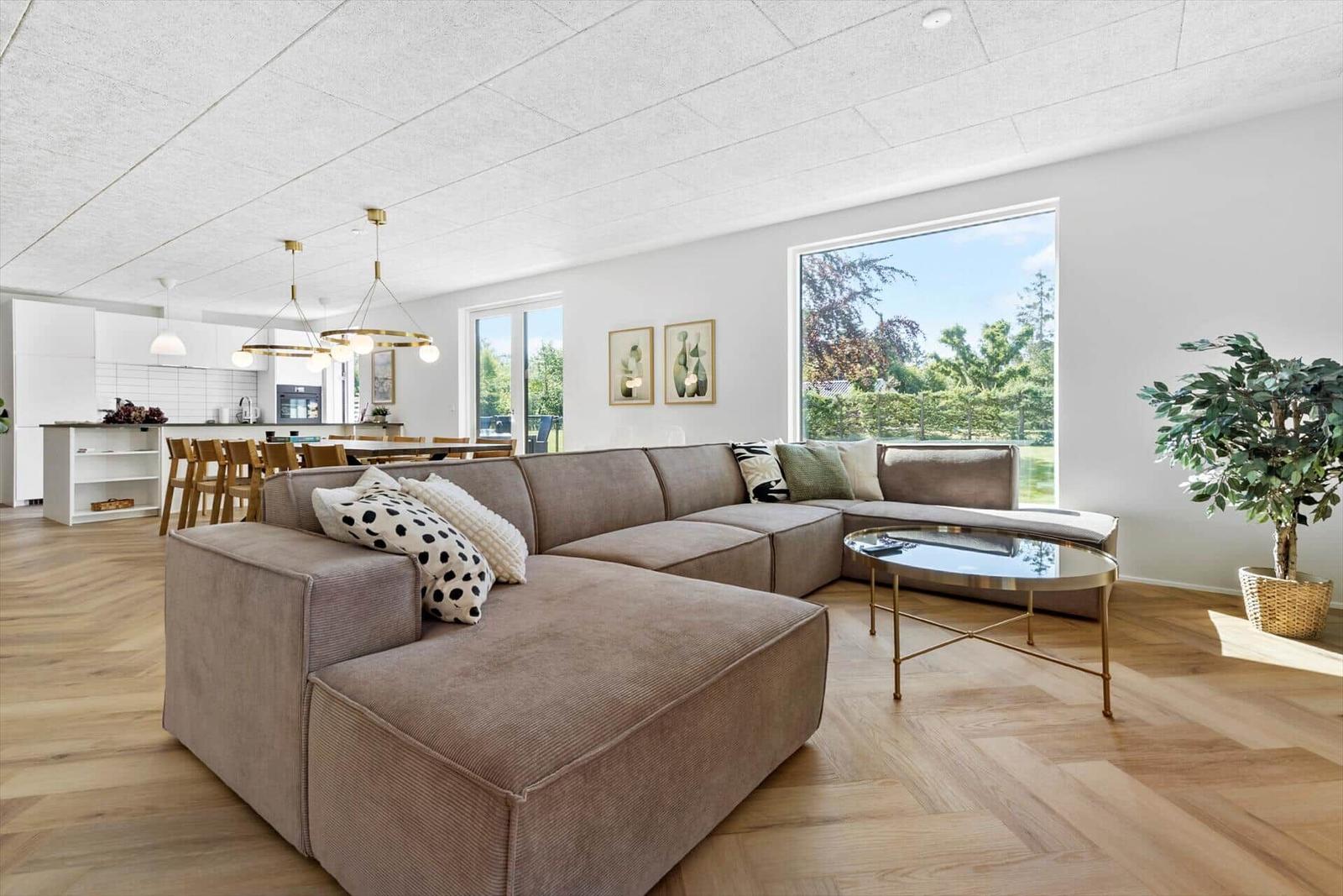 Wohnzimmer mit Ecksofa, Glas-Tisch und Holzboden. Küche im Hintergrund mit Esstisch.