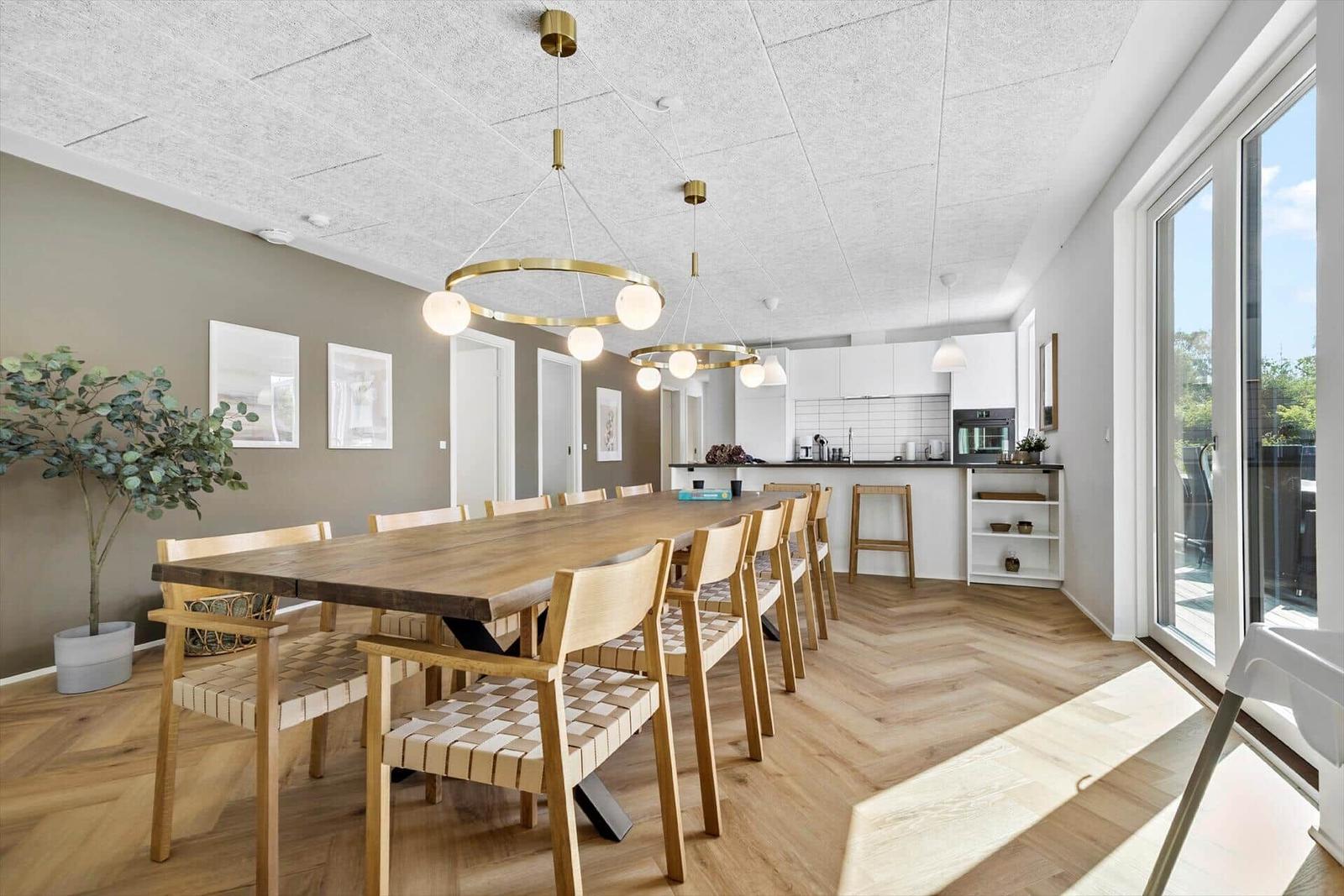 Esszimmer mit langem Holztisch und Stühlen. Küche im Hintergrund. Holzboden und große Fenster.